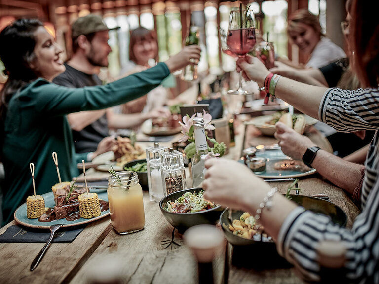 Eine Gruppe Freunde stößt auf das Wiedersehen an einem Tisch im Toni´s Gasthaus, der bereit mit leckeren Grillgerichten gedeckt ist, an.